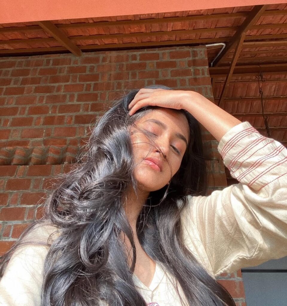Ashlesha Thakur with long wavy hair posing in natural sunlight, eyes closed, with her hand resting on her forehead.