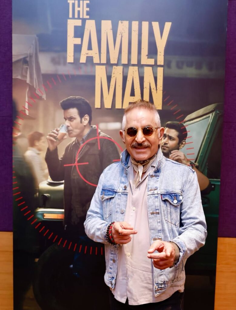 Dalip Tahil wearing sunglasses and a denim jacket posing in front of a promotional poster for “The Family Man"