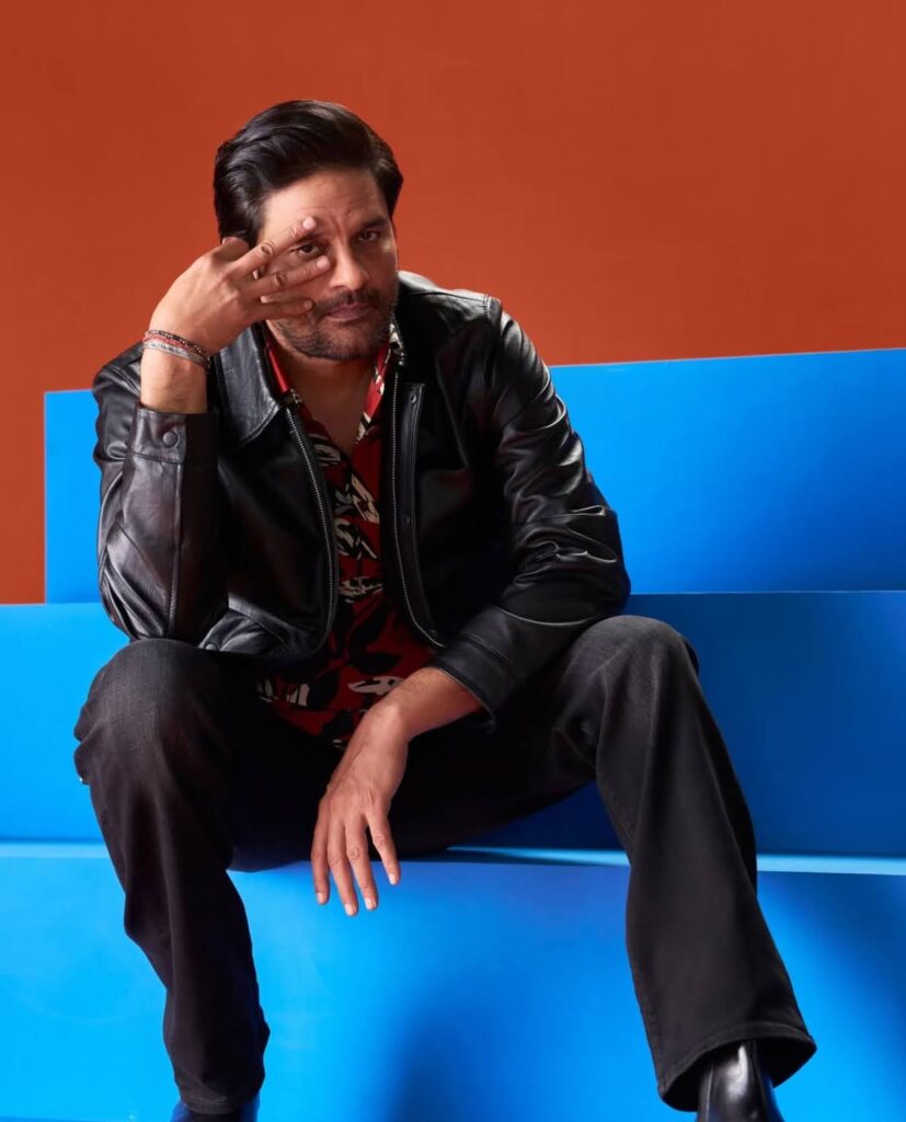 Jaideep Ahlawat in a leather jacket sitting on blue steps with an orange background, making a hand gesture.