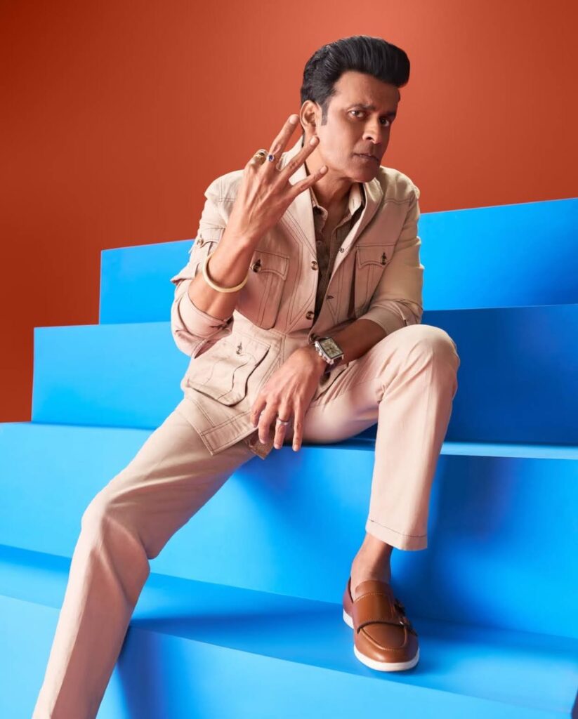 Manoj in a beige outfit sitting on blue steps against an orange backdrop, holding up three fingers.