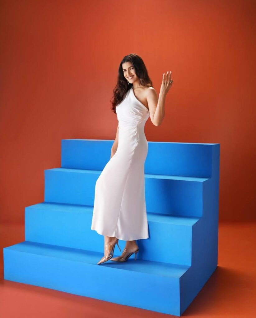 A woman in a white dress standing on blue steps with an orange background, smiling and holding up three fingers.