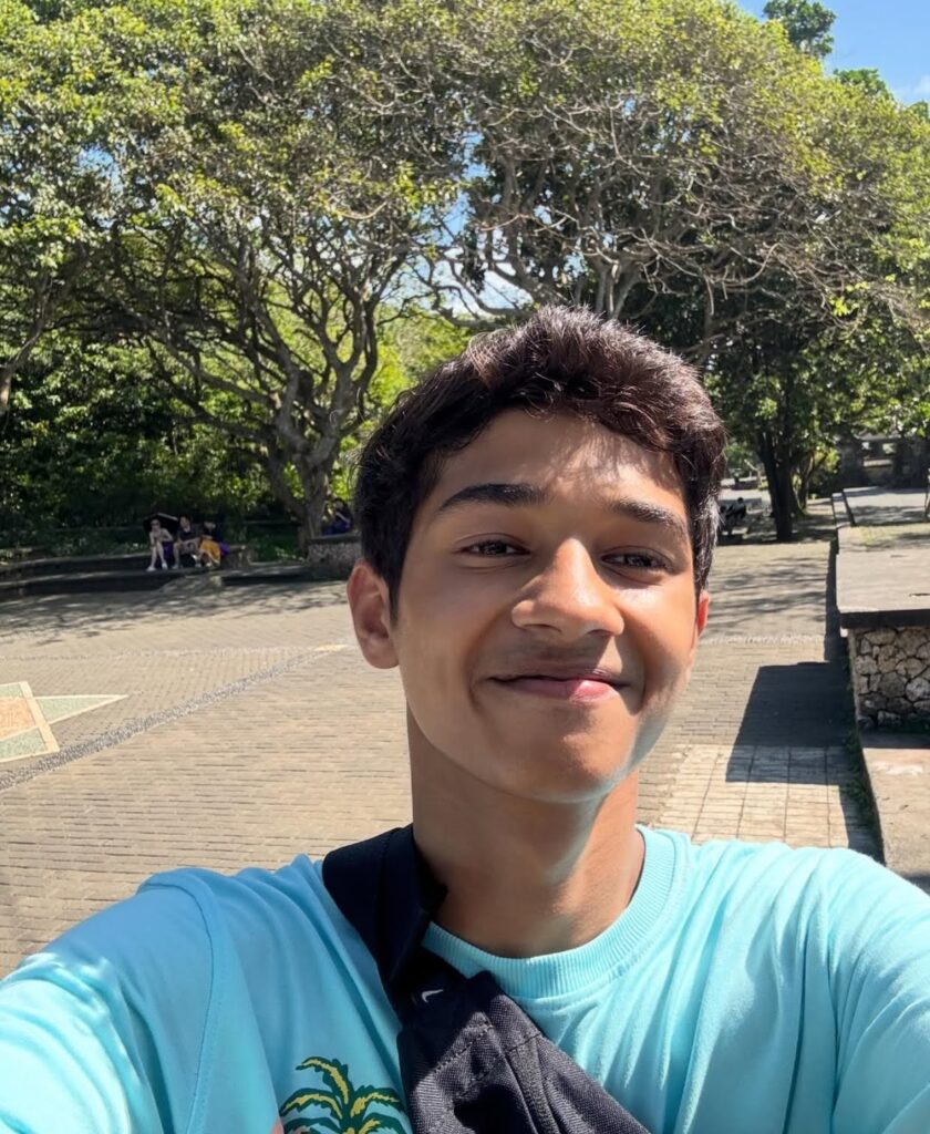 A young man smiling while taking a selfie outdoors, with trees and sunlight in the background.